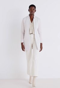 Blazer bianco su una blouse chiara e pantaloni larghi color crema. Tessuto liscio, vestibilità sartoriale e design minimalista.