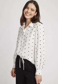Chemise blanche à boutons avec des pois noirs, manches longues avec des poignets élastiques et un ourlet à cordon pour un ajustement modulable.