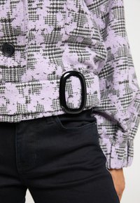 Lilla mønstret jakke med houndstooth-design og sorte accenter, en stor oval sort spænde og tætsiddende mørke jeans.