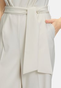 Pantaloni beige chiaro con una cintura in tessuto ampia e con un design drappeggiato, tasche laterali e una texture liscia e fluida.