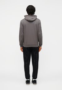 Sivá mikina s kapucňou vo voľnom strihu, kombinovaná s čiernymi vzorovanými teplákmi s bielym nápisom. Modelka viditeľná zozadu.