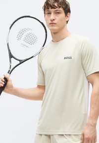 Αθλητική μπλούζα κοντομάνικη, ανοιχτό μπεζ, με στρογγυλή λαιμόκοψη και μαύρο λογότυπο "BOSS", κρατημένη με ρακέτα τένις με μαύρο πλαίσιο και χορδές σε πλέγμα.