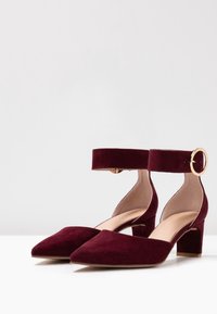 Burgundy mockaskor med klack, spetsig tå och ankelsnodd som har en guldfärgad spänne. Blockklacken ger stabilitet; mjukt innerfoder.