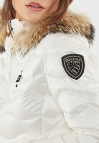 Witte pufferjack met een gequilt ontwerp, voorzien van een met bont gevoerde capuchon en een zwarte patch met strass op de linker mouw.