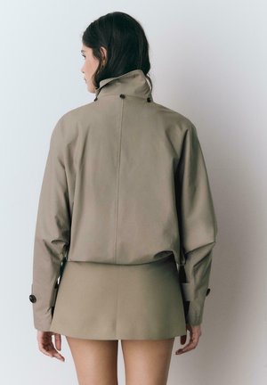 Vrouw met lang donker haar, gekleed in een beige cropped jack en een bijpassende minirok, staand met het gezicht naar een effen lichte achtergrond.