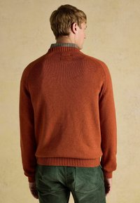 Rostpruuni kudumüts, millel on sooniline krae ja ääre, omades siledat tekstuuri. Kandmiseks ruudulise särk üle, neutraalse tausta ees.