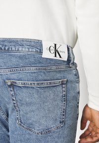Jeans de mezclilla azul claro con una textura suave, que cuentan con una etiqueta blanca cosida en la cintura con texto y logo en negro.