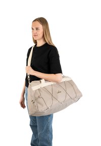 Borsa da viaggio beige con tessuto testurizzato, dettagli con corda elastica e una robusta tracolla color crema, mostrata da una persona su uno sfondo bianco.