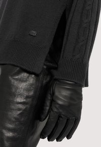 Maglione nero a maglia con spacchi laterali e dettagli a trecce, abbinato a guanti in pelle nera, che mostrano una texture liscia.