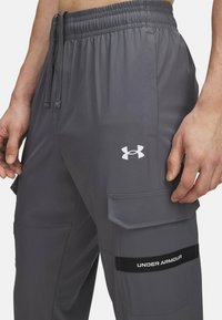 Graue Cargo-Hosen mit einem dehnbaren Bund, ausgestattet mit Seitentaschen und einem weißen Under Armour-Logo am linken Bein. Glatte Textur.