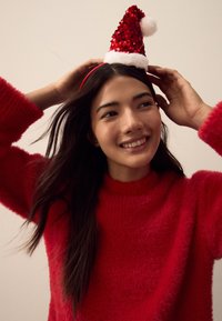 Red fuzzy sweater, hands adjusting a sparkling red and white Santa hat with a pom-pom on top. Simple background enhances the festive look.