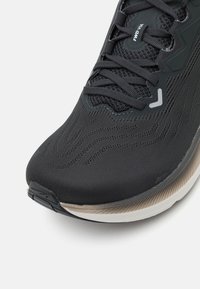 Chaussure de running noire avec tige en mesh texturé, fermeture à lacets, logo de marque proéminent et semelle de couleur claire contrastante avec un motif.