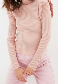 Maglione a collo alto a coste di un rosa chiaro con dettagli a volants sulle spalle e maniche lunghe. Presenta bottoni dorati sui polsini delle maniche.
