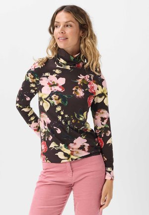 Frau trägt einen schwarzen, floralen Langarm-Rollkragenpullover und pinke Hose, steht mit einer Hand in der Hüfte und blickt zur Seite.