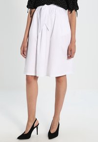 Gonna bianca fino al ginocchio con cintura annodata e design plissettato, realizzata in tessuto leggero. Abbinata a scarpe con tacchi alti nere.