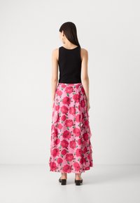 Jupe maxi florale rose avec des volants superposés, ornée de grandes roses rouges et de feuilles vertes, portée avec un débardeur noir et des sandales à talons.