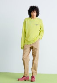 Jeune personne avec des cheveux bouclés, portant un pull vert citron, un pantalon beige et des chaussures rouges, se tenant devant un fond pâle.