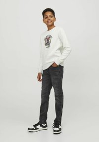 Sudadera gris claro con un diseño gráfico, combinada con unos pantalones vaqueros oscuros. Unas zapatillas de deporte en blanco y negro completan el atuendo.