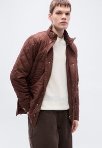 Καφέ υφασμάτινο μπουφάν με όρθιο γιακά, κλείσιμο με φερμουάρ και κουμπώματα με κουμπιά. Φορεμένο πάνω από ένα λευκό πλεκτό πουλόβερ με υφή και σκούρο παντελόνι.