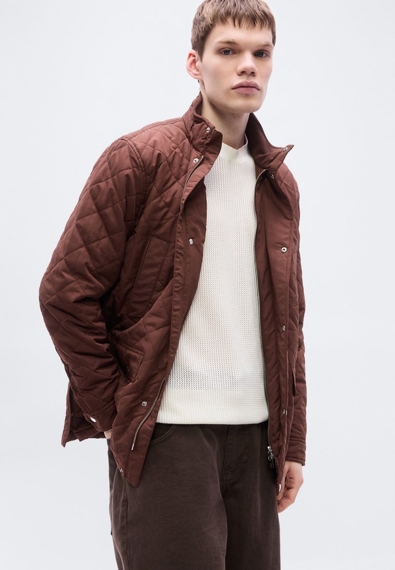 Veste matelassée marron avec col montant, fermeture éclair et boutons-pression. Portée sur un pull blanc texturé et un pantalon foncé.