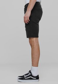 Shorts in denim nero con dettagli strappati, vestibilità slim e orlo grezzo, abbinati a scarpe sportive bianche e nere e calzini bianchi alla caviglia.