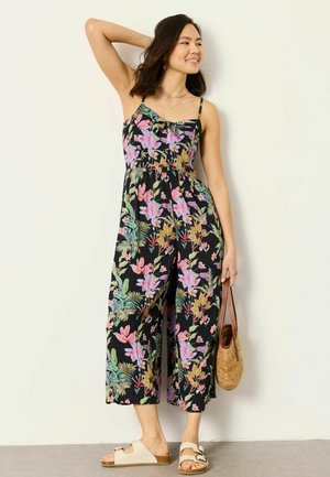 Woman wearing a black floral sleeveless jumpsuit, holding a woven handbag, posing with one arm raised against a plain light wall.