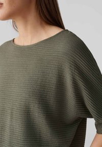 Top verde oliva testurizzato con scollatura ampia, maniche corte a dolman e un morbido motivo a trama, che mette in mostra una vestibilità comoda e rilassata.