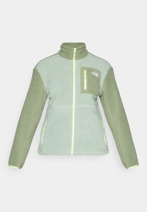 Veste en polaire vert clair avec col haut, devant zippé et manches de couleur vert foncé contrastantes. Comprend une poche poitrine et des poches latérales zippées.