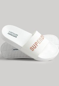 Sandalias blancas de deslizar con una suela texturizada. Presenta una correa ancha con el grabado "SUPERDRY" en dorado rosa. Fabricadas con materiales reciclados.