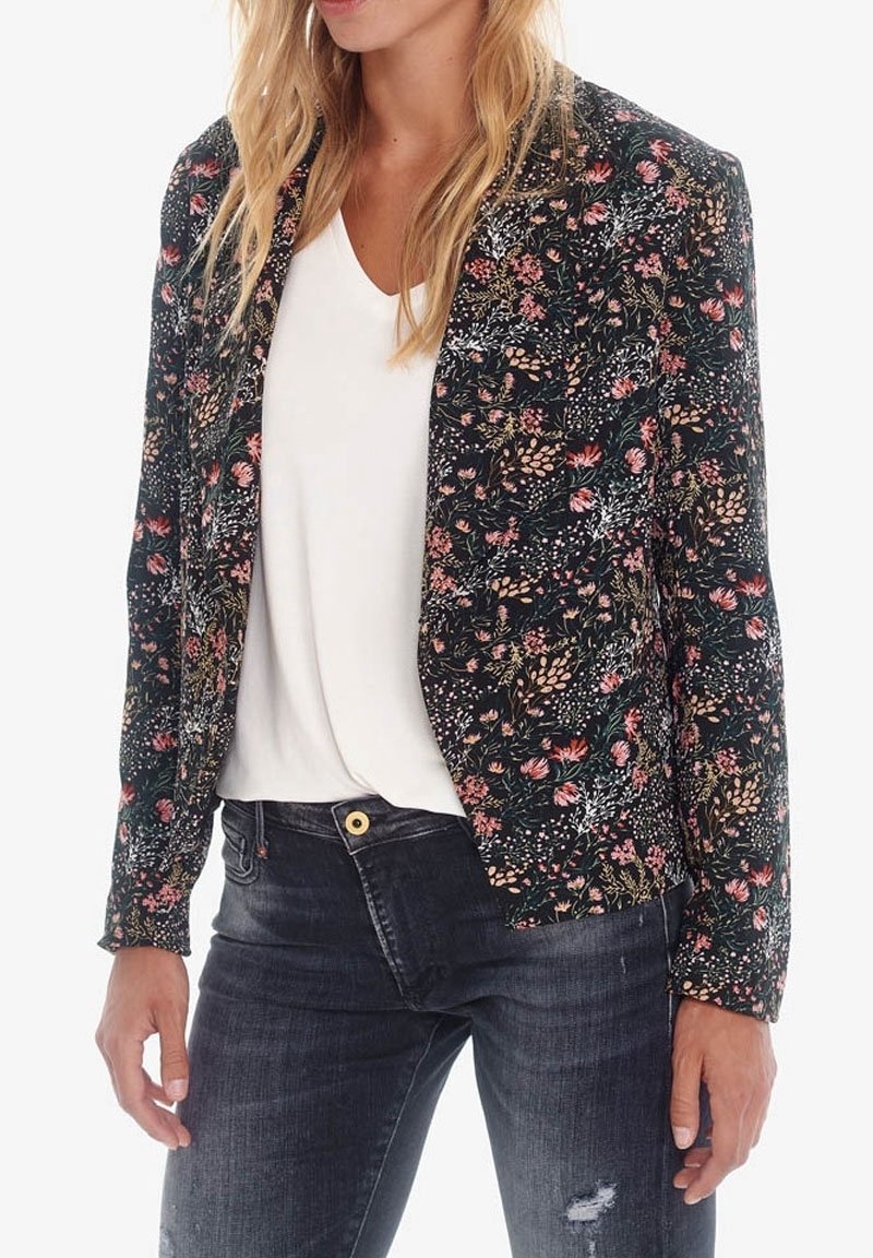 Femme portant un blazer noir fleuri sur un haut blanc et un jean déchiré foncé, debout avec les mains détendues le long du corps.