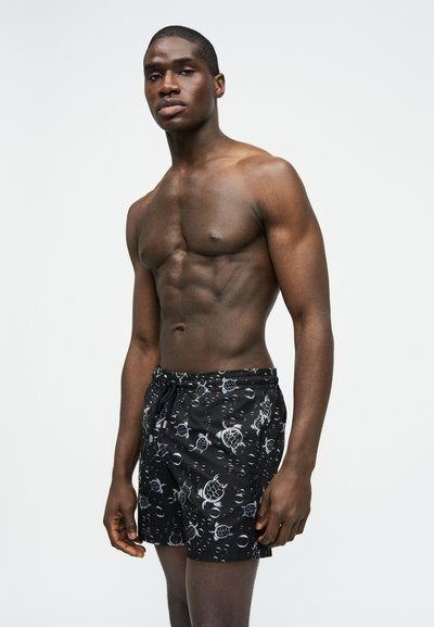 Άνδρας χωρίς πουκάμισο με μυώδη κορμί που φοράει μαύρο μαγιό με λευκά σχέδια θαλάσσιας χελώνας και φυσαλίδων, στέκεται μπροστά σε απλό ανοιχτό φόντο.