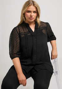 Svart blus med strukturerade prickar, knäppning fram och plisserad design. Har krage och genomskinliga puffärmar.