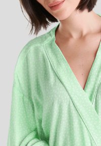 Bata verde claro hecha de tela texturizada con lunares blancos, que cuenta con un cuello de chal y un cierre envolvente.
