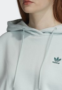 Světle modrá mikina z měkké tkaniny s přední kapsou typu klokan, šňůrkami na kapuci a malým zeleným logem Adidas na prsou.