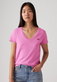 Rožinė V formos marškinėlių su trumpomis rankovėmis, pagaminta iš medvilnės, su mažu Levi's logotipu ant kairiojo krūtinės, derinama su džinsiniais šortais.