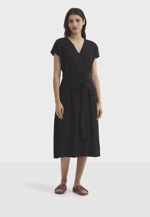 Jeune femme debout portant une robe midi noire à encolure en V, manches courtes et taille nouée, assortie à des sandales rouge à talons ouverts.