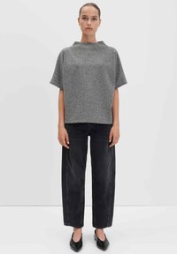 Frau, die vor einem weißen Hintergrund steht und ein graues Kurzarmoberteil, schwarze gerade Jeans und schwarze spitze Ballerinas trägt.