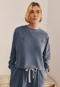 Niebieski bawełniany sweatshirt o luźnym kroju, z ribbowanym dekoltem i wyszywanym logotypem "Wellness". Posiada surowy dół oraz długie rękawy.