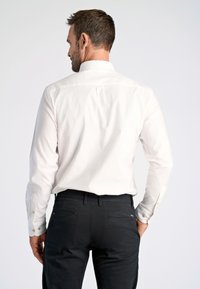 Camicia bianca a maniche lunghe con bottoni, caratterizzata da una sottile texture e un dettaglio con pieghe sul retro, indossata con pantaloni scuri aderenti, che enfatizzano un design pulito.