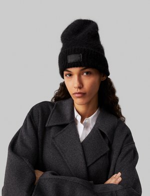 Berretto di lana nera lavorato a maglia con finitura texturizzata, dotato di un'etichetta in pelle con marchio. Il modello indossa un cappotto oversize grigio scuro su una camicia bianca.