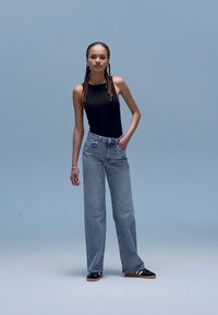 Gaiši pelēkas džinsa bikses ar atslābinātu piegriezumu, kopā ar melnu bezroku topu un melnām slip-on kurpēm. Vienkāršs gaiši zils fons.