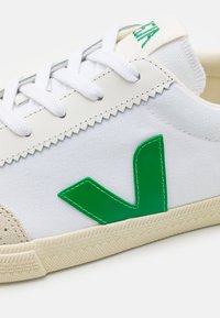 Baltie kedas ar zaļu gumijas V logotipu, auduma augšdaļa, teksturēta bēša pirksta daļa un krēmkrāsas gumijas zole. Iekļautas baltas šņores un šūšanas akcenti.