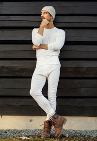 Hvid langærmet skjorte og matchende leggings parret med brune snørebåndsstøvler. Modellen læner sig op ad en sort trævæg og bærer en grå beanie.