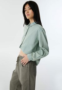 Camicia di jeans cropped blu chiaro con colletto, chiusura a bottoni e maniche lunghe abbinata a pantaloni larghi grigio-verdi con vita elasticizzata.