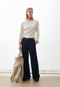 Hellbeiges langärmliges Oberteil, dunkelblaue Jeans mit weitem Bein, strukturierte beige Jacke in der Hand gehalten, dazu schwarze Schuhe und neutraler Hintergrund.