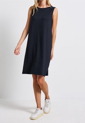 Kobieta w czarnej, bezrękawnikowej sukience do kolan i białych trampkach, stojąca na tle jednolitego, jasnoszarego tła.