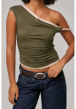 Frau trägt asymmetrisches olivgrünes und beiges Top mit einer freiliegenden Schulter, dunkelblaue Jeans, braunen Gürtel und Schmuck an Händen und Hals.