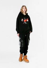 Joven vistiendo una sudadera negra de gran tamaño con un gráfico en rojo y blanco, pantalones cargo negros con cremalleras, y botas de cordones de color beige, apoyado contra un fondo blanco.