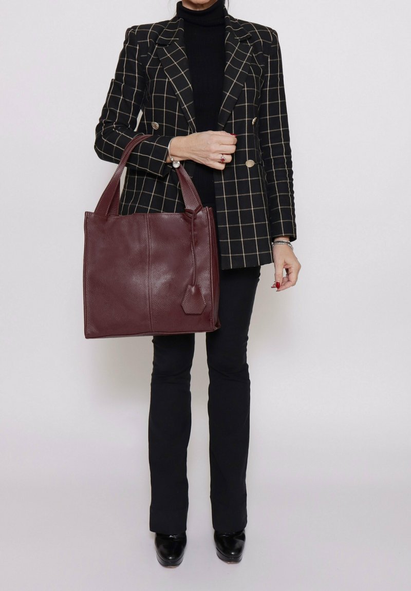 Bordeaux leren handtas met rechthoekige vorm, textuur oppervlak, twee lange handvatten en een labelaccent. Draagt met een geruite blazer en zwarte broek.
