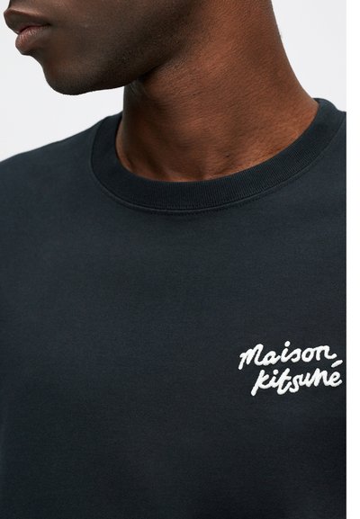 Čierna mikina so štruktúrovaným okrúhlym výstrihom, so žltým výšivkovým logom "Maison Kitsuné" na ľavej strane hrude. Detailný pohľad.